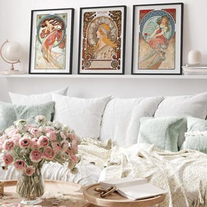 Puede incluir: Tres l&aacute;minas art&iacute;sticas enmarcadas con ilustraciones al estilo de Alphonse Mucha de mujeres con prendas fluidas y fondos circulares complejos, expuestas en una estanter&iacute;a blanca sobre un sof&aacute; con flores rosas en un jarr&oacute;n.