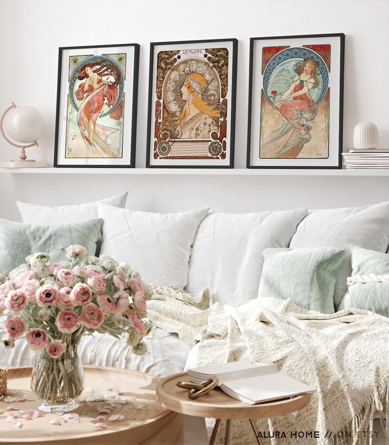 Puede incluir: Tres l&aacute;minas art&iacute;sticas enmarcadas con ilustraciones al estilo de Alphonse Mucha de mujeres con prendas fluidas y fondos circulares complejos, expuestas en una estanter&iacute;a blanca sobre un sof&aacute; con flores rosas en un jarr&oacute;n.