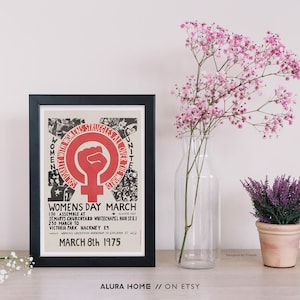 Puede incluir: Un p&oacute;ster enmarcado con borde negro, que muestra un s&iacute;mbolo de pu&ntilde;o rojo y texto sobre una Marcha del D&iacute;a de la Mujer en 1975. Junto a &eacute;l, un jarr&oacute;n de cristal transparente contiene flores rosas y una peque&ntilde;a planta en maceta con flores moradas.