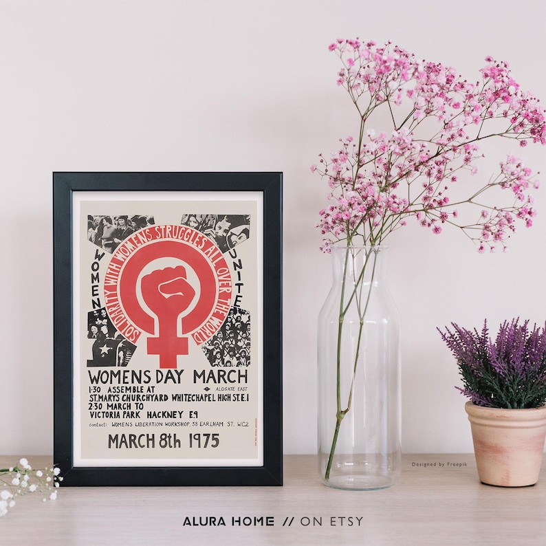 Puede incluir: Un p&oacute;ster enmarcado con borde negro, que muestra un s&iacute;mbolo de pu&ntilde;o rojo y texto sobre una Marcha del D&iacute;a de la Mujer en 1975. Junto a &eacute;l, un jarr&oacute;n de cristal transparente contiene flores rosas y una peque&ntilde;a planta en maceta con flores moradas.