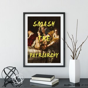 Puede incluir: Una impresi&oacute;n enmarcada en negro de una pintura que representa a tres figuras. Las figuras est&aacute;n en lucha, con una figura sujetando la cabeza de otra. La impresi&oacute;n tiene el texto "SMASH THE PATRIARCHY" en pintura amarilla.