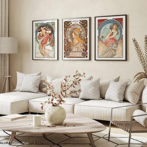 Puede incluir: Un conjunto de tres l&aacute;minas de arte enmarcadas con ilustraciones al estilo de Alphonse Mucha de mujeres con prendas fluidas y bordes decorativos circulares, expuestas en marcos negros sobre una pared encima de un sof&aacute;.
