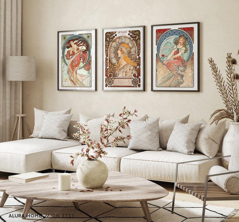 Puede incluir: Un conjunto de tres l&aacute;minas de arte enmarcadas con ilustraciones al estilo de Alphonse Mucha de mujeres con prendas fluidas y bordes decorativos circulares, expuestas en marcos negros sobre una pared encima de un sof&aacute;.