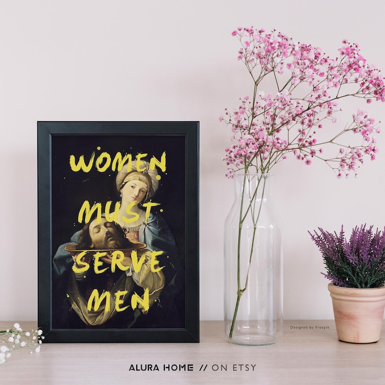 May include: Framed art print with a black frame featuring a Renaissance-style painting. The artwork depicts a woman holding a platter with a head, with the text "WOMEN MUST SERVE MEN" in yellow. A vase of pink flowers and a potted plant are also visible.