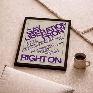 May include: A framed poster with a white background and purple text that reads: GAY LIBERATION FRONT, ARE WE A LOAD OF SCREAMING QUEENS? YES! ARE WE A LOAD OF BUTCH DYKES? YES! FILTHY REDS? YES! COCKSUCKERS? YEAH! FREAKS BEAUTIFUL PEOPLE RIGHT ON.