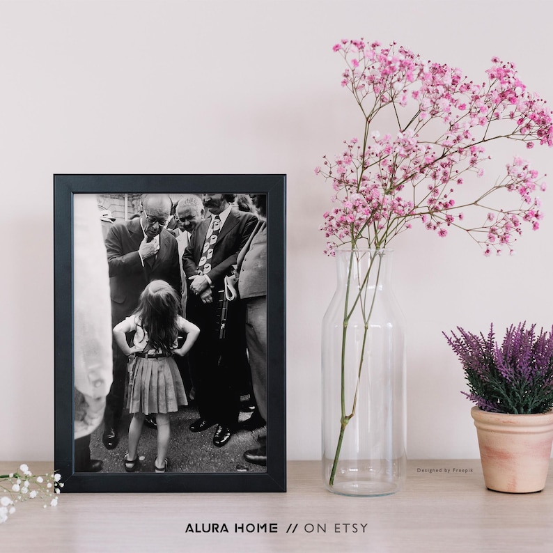 Peut inclure: Une photographie encadr&eacute;e en noir et blanc d'une petite fille devant un groupe d'hommes en costume. Le cadre est noir. Un vase en verre transparent avec des fleurs roses et une petite plante de lavande en pot sont &agrave; droite. Texte en bas : "ALURA HOME // ON ETSY."