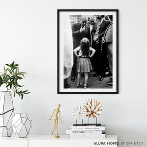 Peut inclure: Tirage encadr&eacute; en noir et blanc repr&eacute;sentant une jeune fille debout, les mains sur les hanches, entour&eacute;e d'hommes en costume. La photographie est de style vintage, capturant un moment dans le temps. L'impression est expos&eacute;e sur une surface blanche.