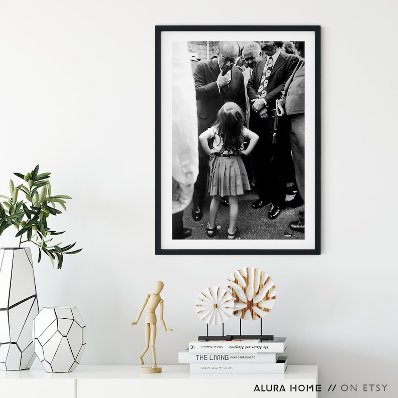 Peut inclure: Tirage encadr&eacute; en noir et blanc repr&eacute;sentant une jeune fille debout, les mains sur les hanches, entour&eacute;e d'hommes en costume. La photographie est de style vintage, capturant un moment dans le temps. L'impression est expos&eacute;e sur une surface blanche.