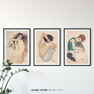 May include: Three framed art prints featuring nude figures in various poses. The prints are in black frames and are hung on a white wall.
