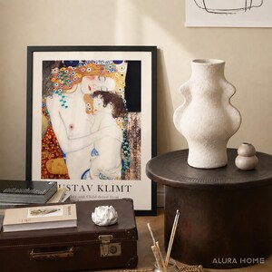 May include: A framed Gustav Klimt Mother and Child art print, a textured off-white ceramic vase with a curved silhouette, a small round beige vase, a brown vintage suitcase, a white lotus-shaped candle holder, and stacked books on a dark table.