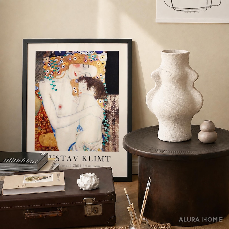 May include: A framed Gustav Klimt Mother and Child art print, a textured off-white ceramic vase with a curved silhouette, a small round beige vase, a brown vintage suitcase, a white lotus-shaped candle holder, and stacked books on a dark table.