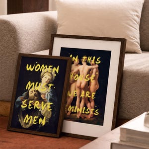 May include: Two framed art prints with dark wood frames. One print features a religious painting with the text "WOMEN MUST SERVE MEN" in yellow. The other print shows figures with the text "IN THIS HOUSE WE ARE FEMINISTS".