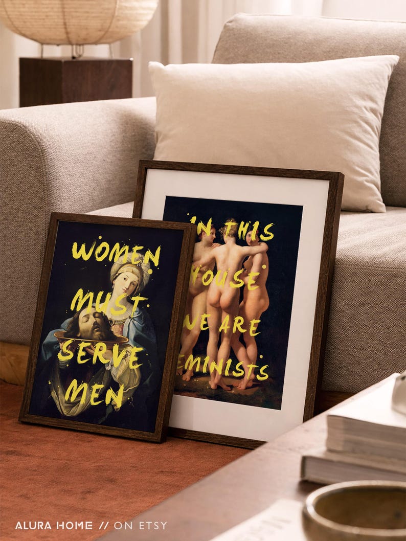 May include: Two framed art prints with dark wood frames. One print features a religious painting with the text "WOMEN MUST SERVE MEN" in yellow. The other print shows figures with the text "IN THIS HOUSE WE ARE FEMINISTS".