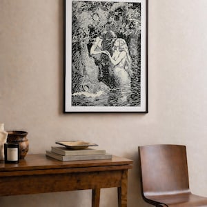 May include: Black and white framed art print depicting two mermaids in a forest scene. The artwork is displayed above a wooden table with books, a decorative bowl, and a candle. A wooden chair is next to the table.