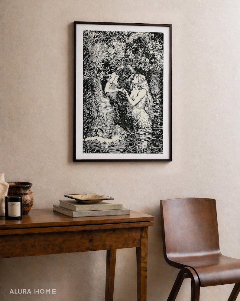 May include: Black and white framed art print depicting two mermaids in a forest scene. The artwork is displayed above a wooden table with books, a decorative bowl, and a candle. A wooden chair is next to the table.
