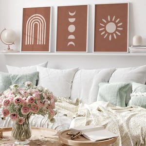 May include: Three framed wall art prints featuring terracotta backgrounds with off-white line art illustrations of a rainbow, moon phases, and a sun. The prints are displayed in white frames on a shelf above a sofa with pillows and a flower vase.