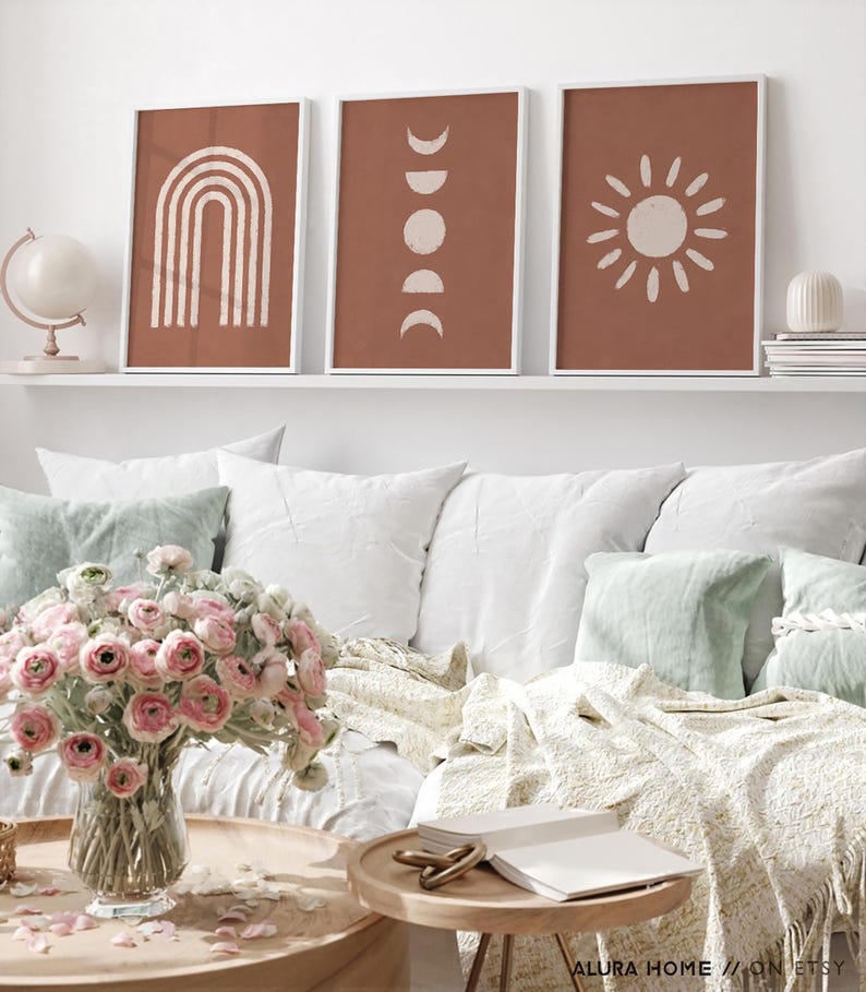 May include: Three framed wall art prints featuring terracotta backgrounds with off-white line art illustrations of a rainbow, moon phases, and a sun. The prints are displayed in white frames on a shelf above a sofa with pillows and a flower vase.