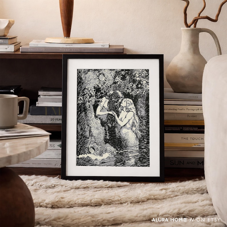May include: Black and white framed art print depicting two figures in a forest setting. One figure is reaching towards the other, who is standing in water. The art print is displayed on a shelf with books and a mug.