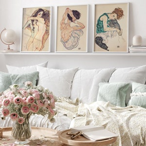 May include: Three framed art prints featuring nude figures in various poses. The prints are in a white frame and are hung on a white wall above a white couch.