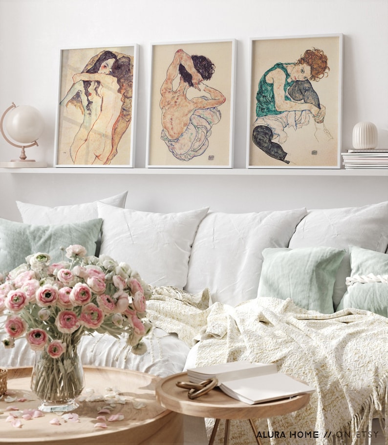 May include: Three framed art prints featuring nude figures in various poses. The prints are in a white frame and are hung on a white wall above a white couch.
