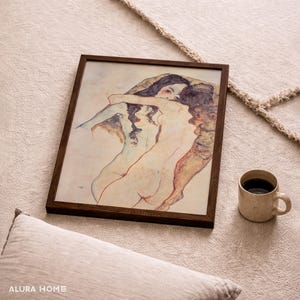 K&ouml;nnte beinhalten: Ein gerahmter Kunstdruck mit einer Aquarell-Illustration von zwei sich umarmenden Figuren. Das Kunstwerk verwendet sanfte Hautt&ouml;ne, ged&auml;mpfte Blaut&ouml;ne und dunkle Haar-Details in einem dunkelbraunen Holzrahmen, pr&auml;sentiert neben einer Tasse.