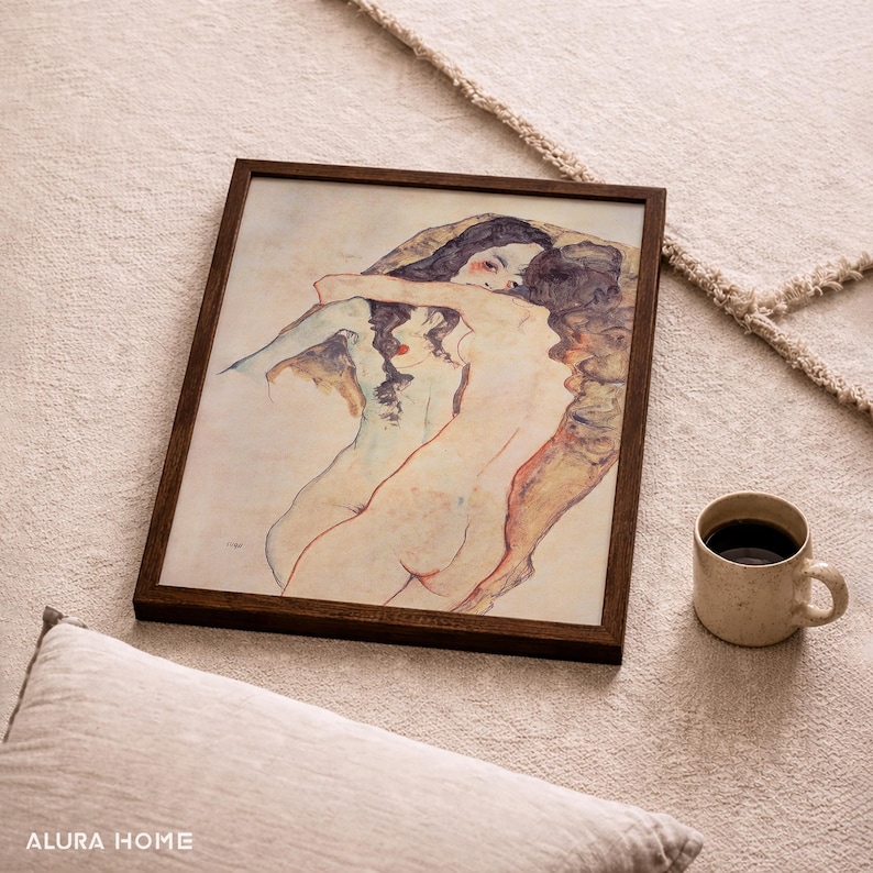 K&ouml;nnte beinhalten: Ein gerahmter Kunstdruck mit einer Aquarell-Illustration von zwei sich umarmenden Figuren. Das Kunstwerk verwendet sanfte Hautt&ouml;ne, ged&auml;mpfte Blaut&ouml;ne und dunkle Haar-Details in einem dunkelbraunen Holzrahmen, pr&auml;sentiert neben einer Tasse.