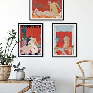 May include: Three framed art prints with a red background and black frames. The prints feature stylised illustrations of figures in various poses. The prints are hung on a white wall with a wooden bench and a chair in the foreground.