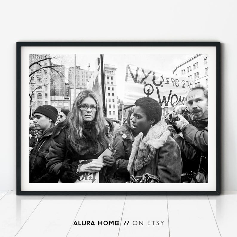 Black and white photograph of Gloria Steinem at a women’s rights protest, surrounded by activists, signs, and photographers in a city square.