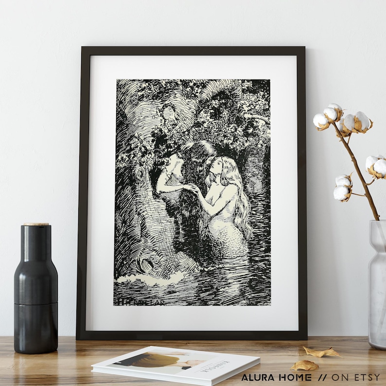 May include: Black and white framed art print depicting two mermaids in a forest setting. One mermaid has long flowing hair and is partially submerged in water. The other mermaid is reaching out. The print is in a black frame.