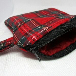 May include: A small, open plaid pouch with a black zip and a red interior. The pouch is red with green, white, and black plaid. The zip is black, and the interior is a solid red colour. The pouch has a small fabric loop.