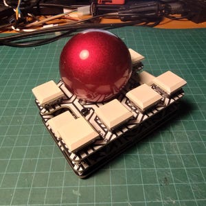 May include: A custom-built electronic device featuring a glossy, deep red sphere atop a black and white patterned circuit board. Square, off-white buttons are arranged around the sphere. The device is on a green grid surface.