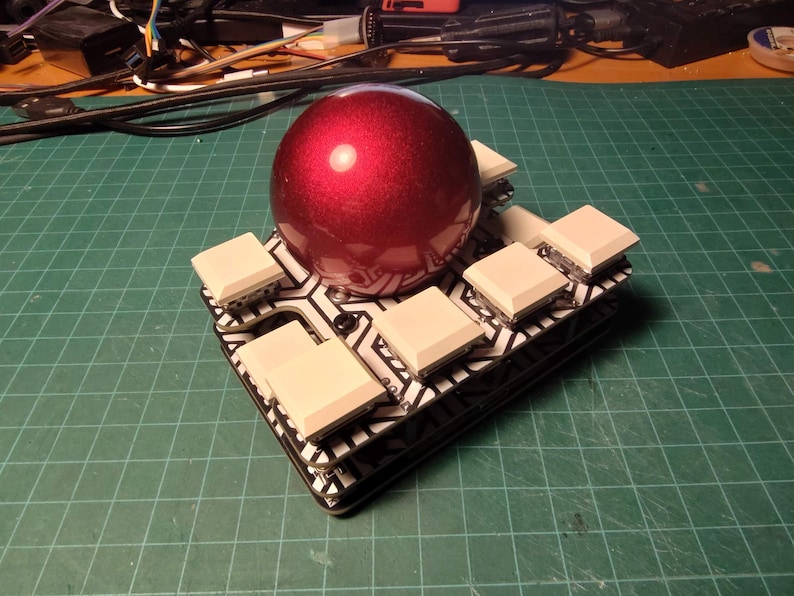 May include: A custom-built electronic device featuring a glossy, deep red sphere atop a black and white patterned circuit board. Square, off-white buttons are arranged around the sphere. The device is on a green grid surface.