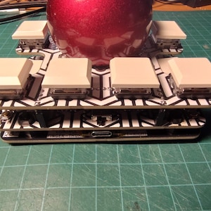 May include: A custom mechanical keyboard with a red, shiny, spherical object on top. The keyboard has white keycaps and a black and white patterned circuit board. The text "FINGER-THUMP" is visible on the bottom layer.
