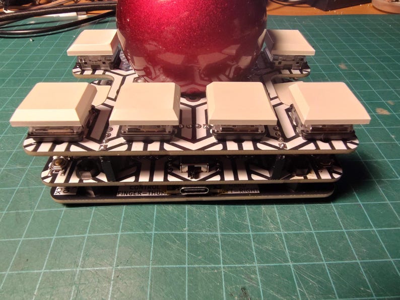 May include: A custom mechanical keyboard with a red, shiny, spherical object on top. The keyboard has white keycaps and a black and white patterned circuit board. The text "FINGER-THUMP" is visible on the bottom layer.