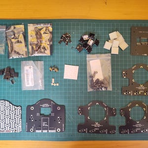 May include: An assortment of electronic components, including circuit boards, connectors, and various small parts, arranged on a green cutting mat. Several components are packaged in clear plastic bags. The layout suggests a DIY electronics project or kit.
