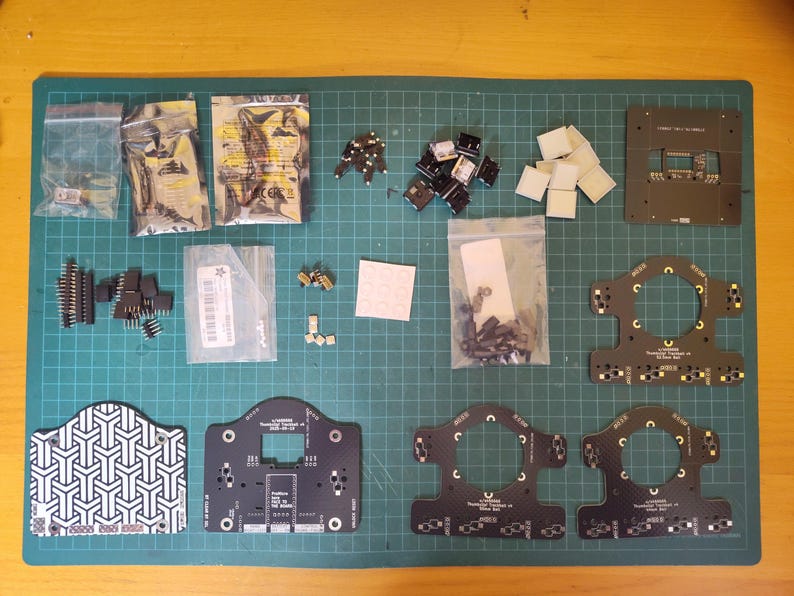May include: An assortment of electronic components, including circuit boards, connectors, and various small parts, arranged on a green cutting mat. Several components are packaged in clear plastic bags. The layout suggests a DIY electronics project or kit.