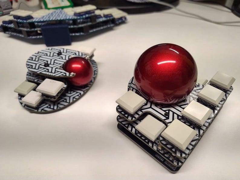 May include: Two custom mechanical keyboard trackballs with a geometric pattern. Each trackball has a glossy red ball and several square, light-colored keys. The keyboards are on a white surface.