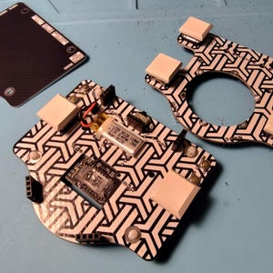 May include: Electronic components arranged on a blue surface. The components include a black and white patterned circuit board, a black rectangular piece, and a white and black patterned frame. The circuit board has a small battery and other electronic parts.