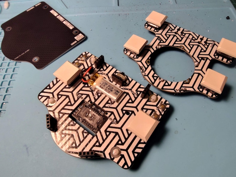 May include: Electronic components arranged on a blue surface. The components include a black and white patterned circuit board, a black rectangular piece, and a white and black patterned frame. The circuit board has a small battery and other electronic parts.
