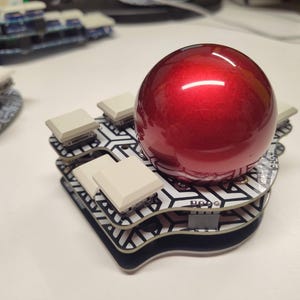 May include: A red, glossy sphere sits atop a custom-built keyboard with square, off-white keycaps. The keyboard has a black and white geometric pattern. The design is modern and unique.