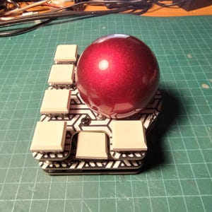 May include: A custom-built mechanical keyboard with a large, shiny, red spherical keycap. The keyboard has a geometric black and white pattern and several square, off-white keycaps. The background is a green grid.