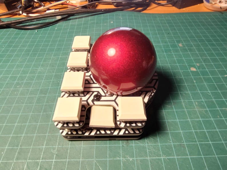 May include: A custom-built mechanical keyboard with a large, shiny, red spherical keycap. The keyboard has a geometric black and white pattern and several square, off-white keycaps. The background is a green grid.