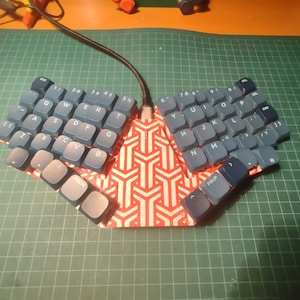 Può includere: Una tastiera blu e bianca con un motivo geometrico rosso e bianco al centro. La tastiera è divisa in due metà, con la metà sinistra che contiene i tasti per le lettere QWERTY e la metà destra che contiene i tasti per le lettere UIO.