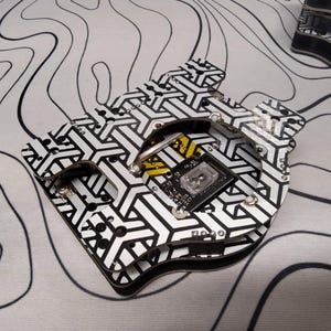 May include: A black and white patterned circuit board with a geometric design. The board has a small black component with text and a clear square component. The background is a white surface with black wavy lines.