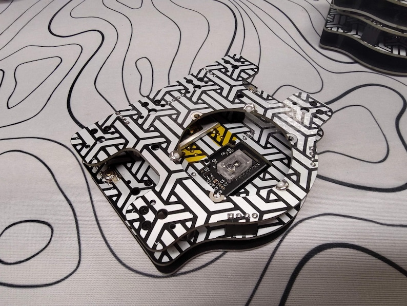 May include: A black and white patterned circuit board with a geometric design. The board has a small black component with text and a clear square component. The background is a white surface with black wavy lines.
