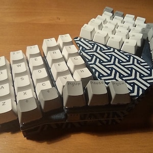 May include: A white mechanical keyboard with a blue and white geometric pattern on the base. The keyboard is split into two sections, with the left section containing the keys for the letters A through Z, and the right section containing the keys for the numbers 1 through 9, the function keys, and the arrow keys. The keyboard also has a number of additional keys, including the Escape key, the Backspace key, the Enter key, the Tab key, the Caps Lock key, the Shift key, the Control key, the Alt key, the Space bar, the Delete key, the Home key, the End key, the Page Up key, the Page Down key, the Insert key, the Print Screen key, the Scroll Lock key, the Pause key, and the Break key.