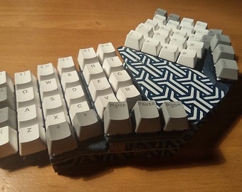 Puede incluir: Un teclado mecánico blanco con un patrón geométrico azul y blanco en la base. El teclado está dividido en dos secciones, la sección izquierda contiene las teclas para las letras A a Z, y la sección derecha contiene las teclas para los números 1 a 9, las teclas de función y las teclas de flecha. El teclado también tiene una serie de teclas adicionales, incluyendo la tecla Escape, la tecla Retroceso, la tecla Intro, la tecla Tabulador, la tecla Bloq Mayús, la tecla Mayús, la tecla Control, la tecla Alt, la barra espaciadora, la tecla Eliminar, la tecla Inicio, la tecla Fin, la tecla Re Pág, la tecla Av Pág, la tecla Insertar, la tecla Impr Pant, la tecla Bloq Despl, la tecla Pausa y la tecla Interrupción.