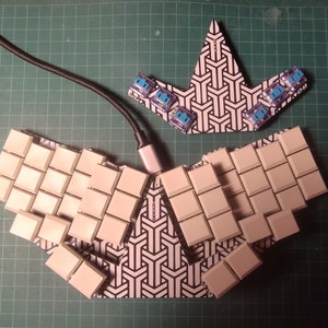 Puede incluir: Un teclado ergonómico dividido con teclas blanquecinas e interruptores azules. El teclado está conectado a un cable negro. La base tiene un patrón geométrico en blanco y negro. El teclado está sobre una esterilla de corte verde.