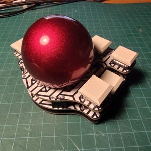 May include: A deep red, spherical object rests on a black and white patterned base, surrounded by several square, off-white blocks. The base has a unique, curved shape. The background is a green grid.
