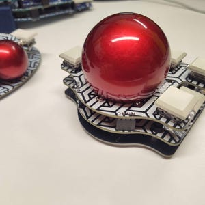 May include: A red, glossy sphere sits atop a layered, geometric patterned base with white square buttons. The base has a black border. Another similar object is visible in the background. The objects are on a white surface.